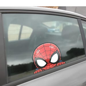 May include: A red and black Spider-Man car window decal. The decal features the character's head and hands, peeking over the edge of the window. The background shows the interior of the car and the outside environment.
