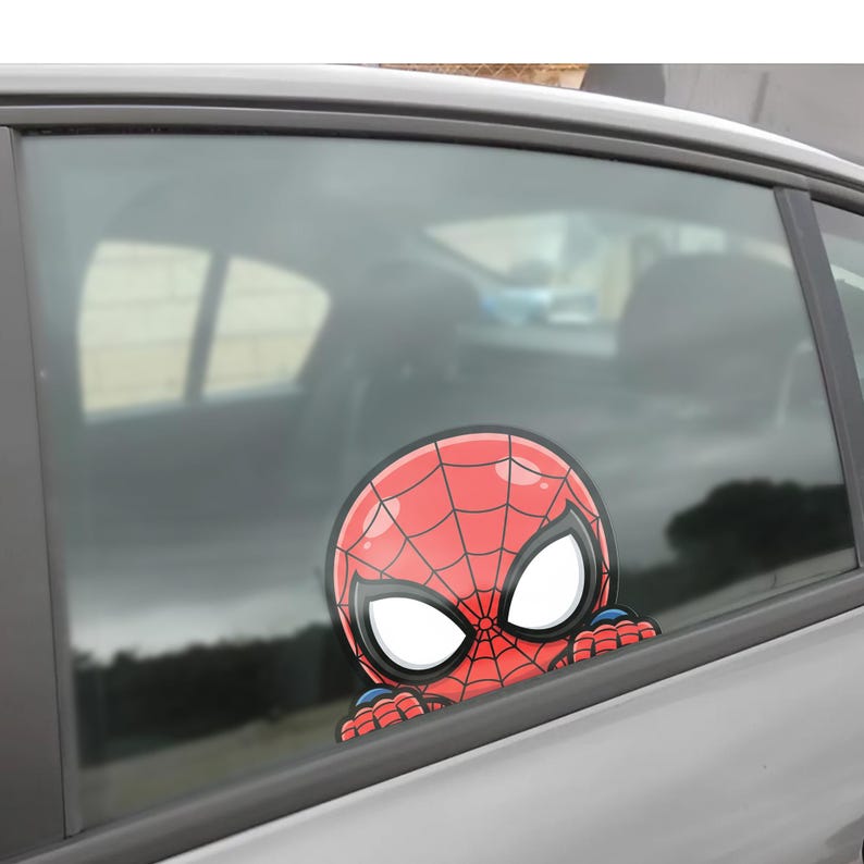 May include: A red and black Spider-Man car window decal. The decal features the character's head and hands, peeking over the edge of the window. The background shows the interior of the car and the outside environment.