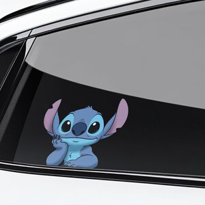 May include: A blue cartoon character sticker, featuring large eyes and large ears, is affixed to a car window. The character is smiling and resting its chin on its hand. The sticker is a fun and whimsical car accessory.
