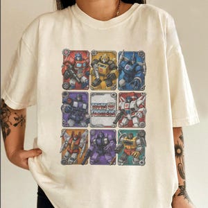May include: A cream-colored short-sleeve t-shirt featuring a graphic print of eight Transformers characters arranged in a grid around a central Transformers logo. The characters are depicted in various colors including red, yellow, blue, and purple.