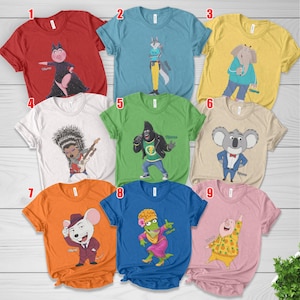 May include: A collection of nine short-sleeved t-shirts in various colors, each featuring a different cartoon character from the movie Sing. The shirts display characters like a pig, wolf, elephant, porcupine, gorilla, koala, mouse, lizard, and piglet, with the word Name printed on each.
