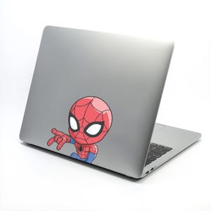 Pode incluir: Um adesivo de desenho animado do Homem-Aranha, em vermelho e azul, est&aacute; afixado na parte de tr&aacute;s de um laptop prateado. O adesivo apresenta a m&aacute;scara do Homem-Aranha com grandes olhos brancos e uma m&atilde;o estendida.