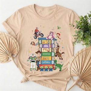 Może przedstawiać: Beżowa koszulka z nadrukiem stosu książek z filmami Disneya z postaciami z Toy Story. Książki zawierają tytuły takie jak "Toy Story", "Piękna i Bestia" i "Kr&oacute;l Lew". R&oacute;żne postacie z kresk&oacute;wek są przedstawione na i wok&oacute;ł książek.