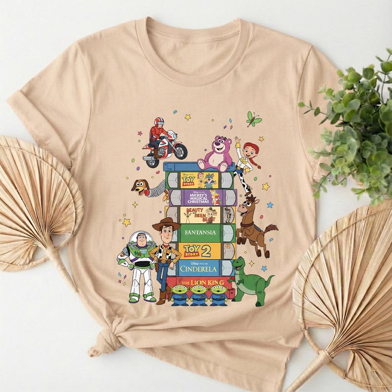 Może przedstawiać: Beżowa koszulka z nadrukiem stosu książek z filmami Disneya z postaciami z Toy Story. Książki zawierają tytuły takie jak "Toy Story", "Piękna i Bestia" i "Kr&oacute;l Lew". R&oacute;żne postacie z kresk&oacute;wek są przedstawione na i wok&oacute;ł książek.