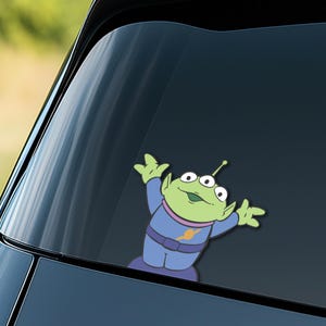 May include: A green alien sticker with three eyes, wearing a blue uniform with a purple belt and collar. The alien is smiling and has its arms raised. The sticker is on a car window.