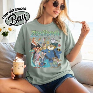 May include: A sage green short-sleeve t-shirt featuring a graphic print of Zootopia characters including Judy Hopps and Nick Wilde. The text Zootopia is printed above the characters. The shirt has a Comfort Colors Bay label in the upper left corner.