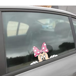 May include: A Minnie Mouse car window decal. The decal features Minnie's face, black ears, and a pink bow with white polka dots. Minnie's white gloved hands are visible, as if she is peeking over the window.