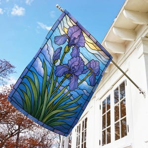 May include: A rectangular garden flag featuring a stained-glass design of purple iris flowers against a blue sky background. The flag is mounted on a metal pole and displayed outside a white house.