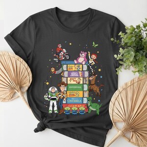 Może przedstawiać: Ciemnoszara koszulka z motywem postaci i tytuł&oacute;w film&oacute;w Disneya na stosie książek. Przedstawiono postacie z Toy Story, Kopciuszka i Kr&oacute;la Lwa. Koszulka ma okrągły dekolt i kr&oacute;tkie rękawy.