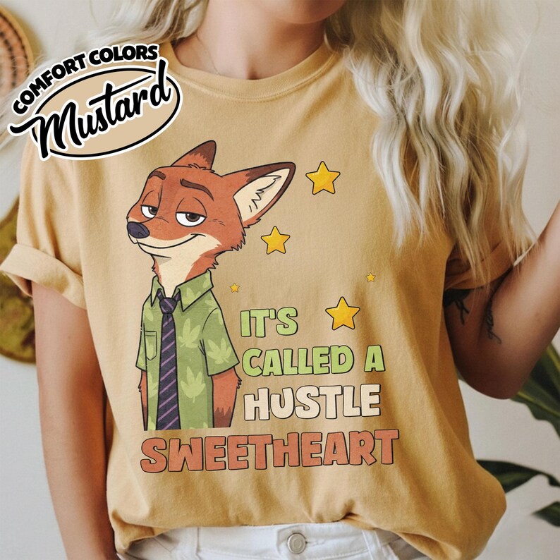 Puede incluir: Una camiseta color mostaza con el gr&aacute;fico de un zorro de dibujos animados que viste una camisa verde con estampado de hojas y una corbata morada. El texto dice IT'S CALLED A HUSTLE SWEETHEART, junto a estrellas amarillas. Incluye logo Comfort Colors.