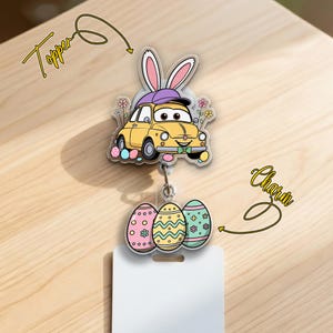 May include: A yellow car-shaped badge reel topper with bunny ears and a purple hat, paired with a charm of three decorated Easter eggs. The topper and charm are attached to a white badge holder.