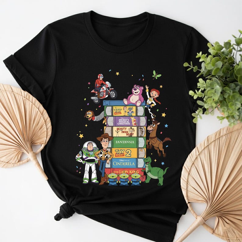 Może przedstawiać: Czarny t-shirt z kolorową grafiką przedstawiającą ułożone w stos książki z tytułami film&oacute;w Disneya i postaciami takimi jak Buzz Lightyear i Woody. Projekt zawiera motocykl, r&oacute;żowego misia i zielonego dinozaura.