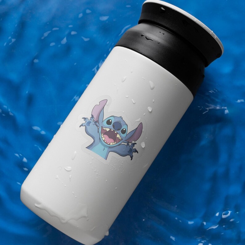 May include: A white insulated tumbler with a black lid, featuring a colorful sticker of the character Stitch from the movie Lilo & Stitch. The sticker shows Stitch with an open mouth and arms outstretched. The tumbler is on a blue surface.