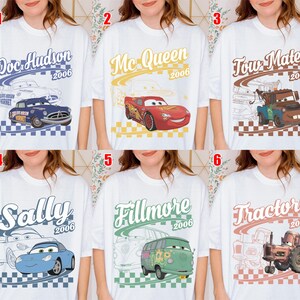 May include: A white t-shirt featuring six distinct designs of characters from the movie Cars, including Doc Hudson, Mc Queen, Tow Mater, Sally, Fillmore, and Tractors, each with their name, the year 2006, and a checkered pattern background.