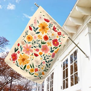 May include: A cream-colored flag with a floral pattern featuring pink, red, yellow, and green flowers. The flag is hanging from a pole against a blue sky and a white house.