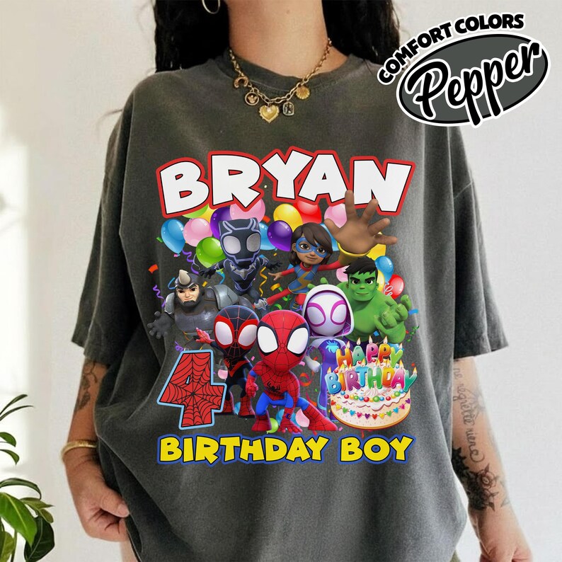 May include: A charcoal gray t-shirt featuring a graphic print with the text BRYAN and BIRTHDAY BOY. The design includes various superhero characters, colorful balloons, a large red number 4, and a birthday cake with lit candles.