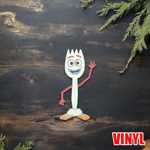 May include: A white and blue cartoon character resembling a spork, with red arms and feet, and a smiling face. The character is waving. The word "VINYL" is in red block letters in the bottom right corner. The background is a dark wood surface.