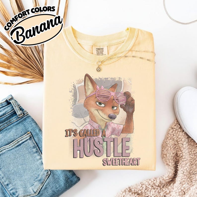 Puede incluir: Una camiseta de cuello redondo color amarillo p&aacute;lido con un gr&aacute;fico de un zorro de dibujos animados que lleva gafas de sol de persiana rosas y una camisa estampada. El texto bajo el zorro dice "IT'S CALLED A HUSTLE SWEETHEART". Etiqueta en el cuello.