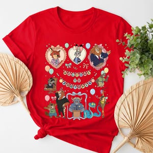 K&ouml;nnte beinhalten: Ein rotes T-Shirt mit einem farbenfrohen Aufdruck von Charakteren aus dem Animationsfilm Zootopia. Das Design enth&auml;lt den Text "WELCOME TO ZOOTOPIA" und verschiedene Tierfiguren in herzf&ouml;rmigen Rahmen.