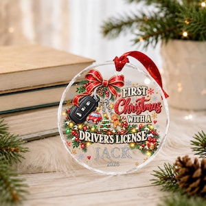 May include: A round, clear acrylic Christmas ornament featuring a red ribbon hanger. The design includes a black car key fob, a red car, a Christmas tree, a gingerbread man, and the text: First Christmas with a Driver's License, Jack, 2026.