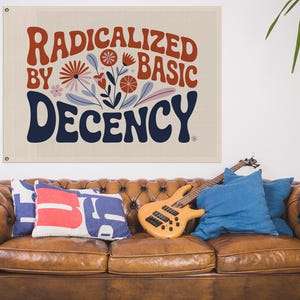 May include: A canvas wall hanging with the words "RADICALIZED BY BASIC DECENCY" in red and navy blue, with floral accents. The wall hanging is above a brown leather sofa with cushions and a guitar.