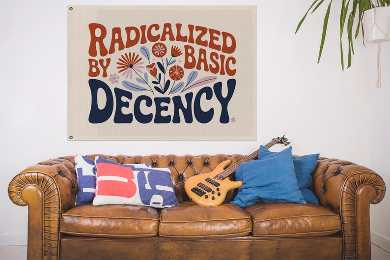May include: A canvas wall hanging with the words "RADICALIZED BY BASIC DECENCY" in red and navy blue, with floral accents. The wall hanging is above a brown leather sofa with cushions and a guitar.