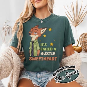 Puede incluir: Una camiseta de cuello redondo en color verde azulado oscuro con un gr&aacute;fico de un zorro de dibujos animados con camisa verde y corbata a rayas. El texto dice IT'S CALLED A HUSTLE SWEETHEART, junto a peque&ntilde;as estrellas amarillas. Corte relajado.