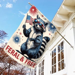 May include: A rectangular flag featuring a cartoon raccoon wearing sunglasses and holding a small American flag. The flag has the text "FERAL & FREE" in red. Fireworks and stars are in the background. The flag is hanging on a white building.
