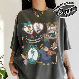 May include: A charcoal gray short-sleeve t-shirt featuring a graphic print of Zootopia characters including Nick Wilde, Judy Hopps, and Flash. The design includes ornate frames, a banner with the text Welcome to Zootopia, and small carrot icons.