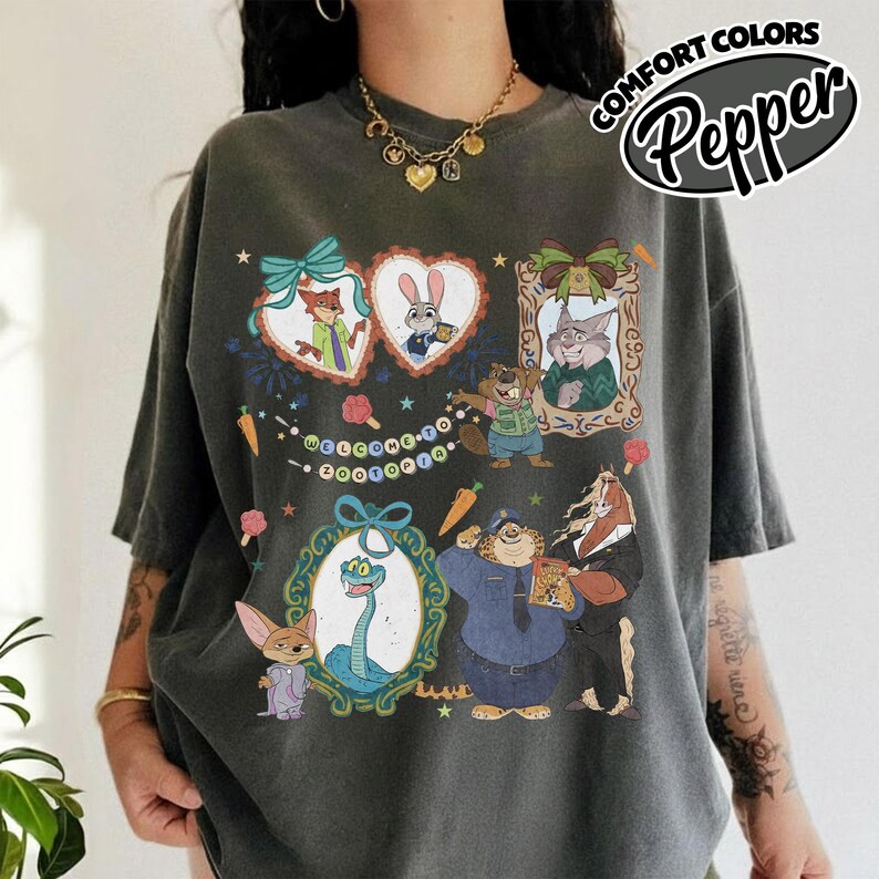 May include: A charcoal gray short-sleeve t-shirt featuring a graphic print of Zootopia characters including Nick Wilde, Judy Hopps, and Flash. The design includes ornate frames, a banner with the text Welcome to Zootopia, and small carrot icons.