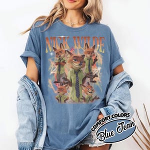May include: A blue jean colored t-shirt featuring a graphic print of the character Nick Wilde in various poses. The text NICK WILDE appears in orange at the top, surrounded by yellow lightning bolt illustrations against a faded blue background.