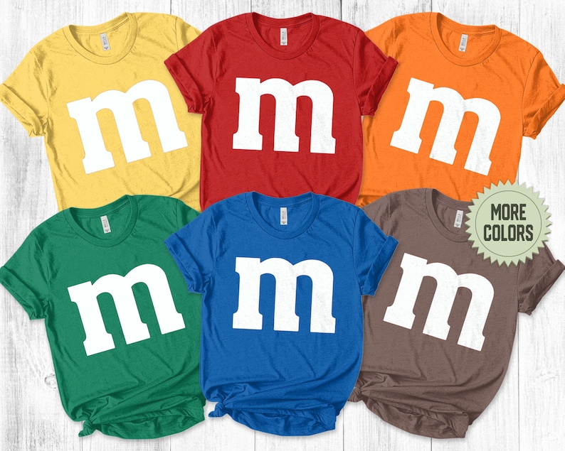 May include: A collection of six short-sleeved crew neck t-shirts in yellow, red, orange, green, blue, and brown. Each shirt features a large, white, lowercase letter m printed on the front center. A circular badge with the text MORE COLORS is included.
