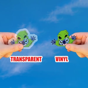 May include: Two stickers featuring a green cartoon character with blue accents, held against a blue sky. One sticker is transparent, the other is vinyl. The word "TRANSPARENT" is in red text below the transparent sticker, and "VINYL" is below the other.