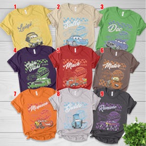 May include: Nine colorful t-shirts featuring characters from the movie Cars. Each shirt has a different character's name and image, including Luigi, Sally, Doc, Chick Hicks, Mack, Mater, McQueen, Guido, and Ramone. The shirts are short-sleeved.