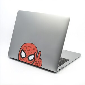 Pu&ograve; includere: Un laptop argentato con un adesivo di Spider-Man rosso e nero sul retro. L'adesivo mostra la maschera di Spider-Man con occhi bianchi e una mano che fa il segno della pace.