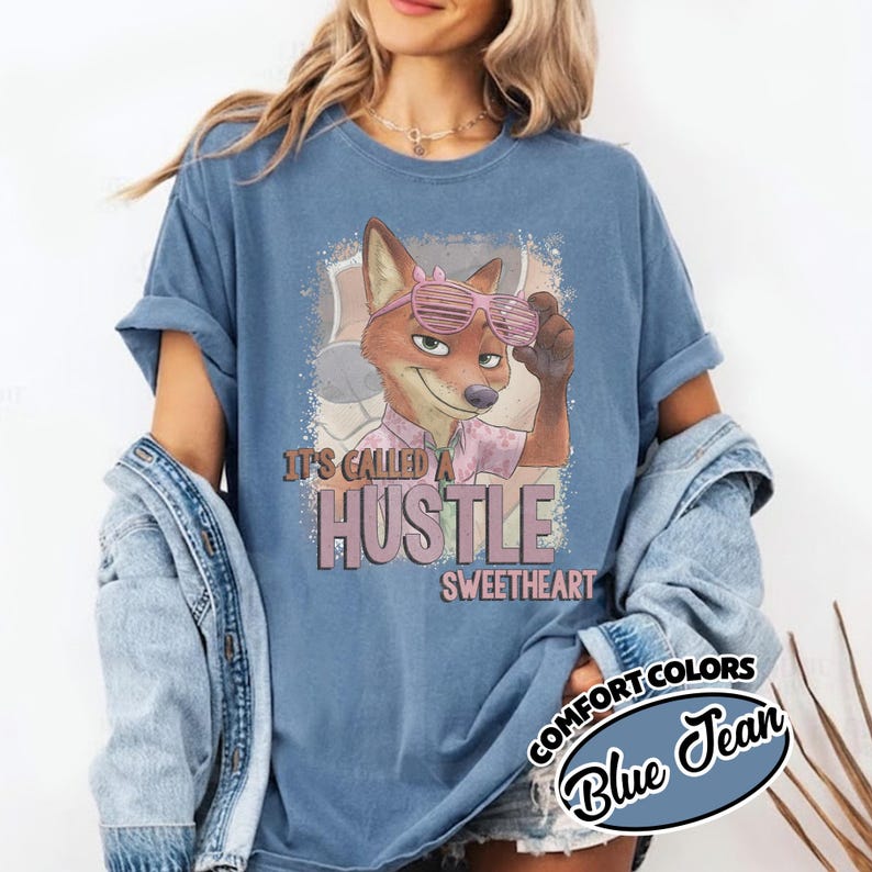 Puede incluir: Una camiseta de manga corta color azul jean con un gr&aacute;fico de un zorro de dibujos animados que lleva gafas de sol de persiana rosas. El texto en la camiseta dice, It's called a hustle sweetheart. Tiene cuello redondo y un corte holgado.