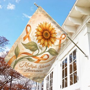 May include: A decorative flag featuring a large sunflower with orange ribbons and green leaves. The flag has the words "Multiple Sclerosis Awareness" in a script font. The flag is hanging on a white house.