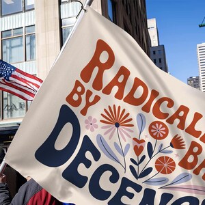 May include: A cream flag with the words "RADICALIZED BY DECENCY" in orange and navy blue, with floral illustrations. An American flag waves in the background, with a city skyline and blue sky.