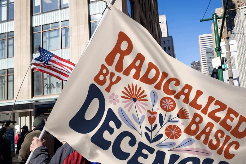May include: A cream flag with the words "RADICALIZED BY DECENCY" in orange and navy blue, with floral illustrations. An American flag waves in the background, with a city skyline and blue sky.