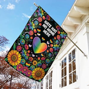 May include: A black flag with a floral design and the text "Hate Has No Home Here." The flag features a rainbow heart and smaller rainbow hearts, along with colorful flowers, including sunflowers, and leaves. The flag is hanging outside a white building.