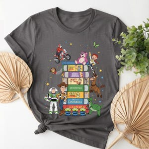 Może przedstawiać: Ciemnoszara koszulka z kolorową grafiką postaci z kresk&oacute;wek z Toy Story i innych film&oacute;w Disneya. Projekt zawiera postacie takie jak Buzz Lightyear i Woody, ułożone na stosie kaset VHS.
