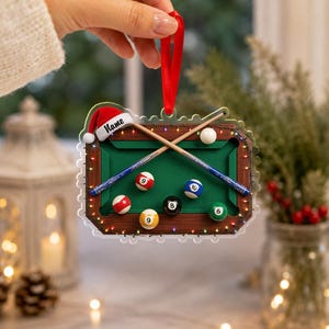May include: A rectangular acrylic Christmas ornament featuring a green pool table with brown wooden rails, two crossed pool cues, and six billiard balls numbered 6, 8, 9, 9, and 10. A red Santa hat sits on the top left corner with the text Name.