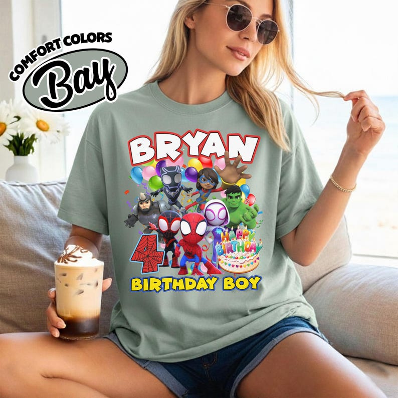 May include: A sage green t-shirt featuring a graphic print of various superhero characters, including Spider-Man, Hulk, and Black Panther. The design includes the text "BRYAN", "4", and "BIRTHDAY BOY" along with a birthday cake and colorful balloons.