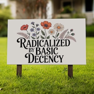 May include: A rectangular sign with the text "RADICALIZED BY BASIC DECENCY" in black script. The sign features a floral design with red, orange, blue, and white flowers. The sign is mounted on two wooden stakes and set in a grassy yard.