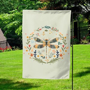 May include: A cream-colored garden flag featuring a detailed dragonfly design. The dragonfly is centered within a floral wreath with orange, yellow, and blue accents. The flag is hanging on a black metal stand in a grassy yard.