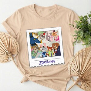 May include: A beige t-shirt featuring a graphic of characters from the animated film "Zootopia" in a polaroid-style frame. The frame includes the film's title. The shirt is made of a soft material.