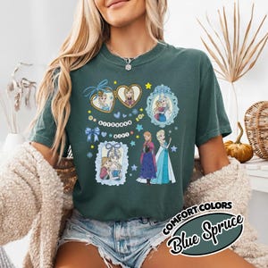 May include: A dark green short-sleeve t-shirt featuring a graphic print of two animated characters in heart-shaped and ornate frames, along with the text "BIRTHDAY GIRL" on a string of beads, surrounded by blue bows, stars, and snowflakes.
