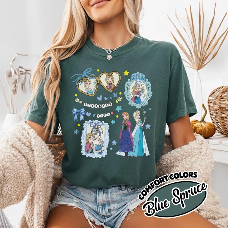 May include: A dark green short-sleeve t-shirt featuring a graphic print of two animated characters in heart-shaped and ornate frames, along with the text "BIRTHDAY GIRL" on a string of beads, surrounded by blue bows, stars, and snowflakes.