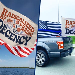 May include: A white flag with the text "RADICALIZED BY BASIC DECENCY" in brown and navy blue, with floral accents. The flag is displayed on a boat and a truck, alongside an American flag.