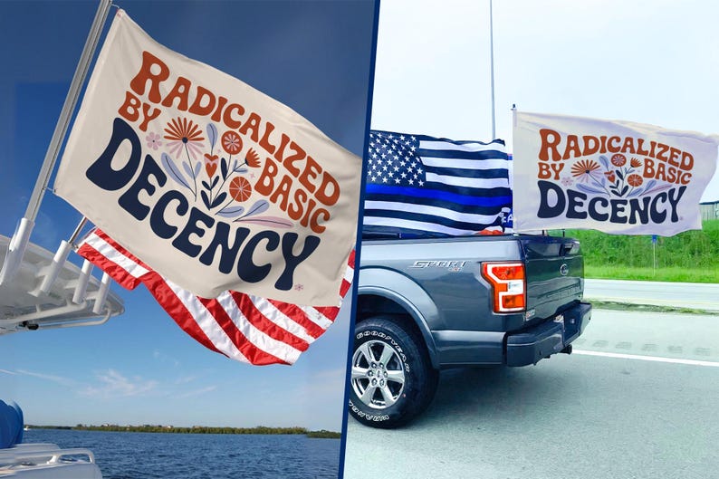 May include: A white flag with the text "RADICALIZED BY BASIC DECENCY" in brown and navy blue, with floral accents. The flag is displayed on a boat and a truck, alongside an American flag.